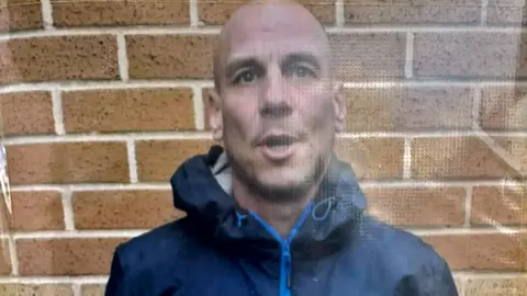 A bald man, who is wearing a dark blue coat with the hood down, has his mouth partially open as he stands in front of a wall.