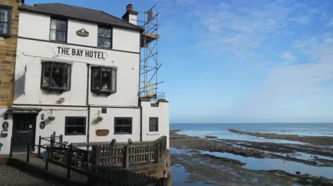 A white seaside building labelled “The Bay Hotel” stands on a cobbled slope beside the shoreline. The building has multiple windows, a doorway to a bar, and scaffolding on its right side. A wooden fence runs along the front. To the right, the rocky beach is exposed at low tide, with shallow pools of water leading out to the calm sea beneath a blue, lightly clouded sky.