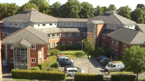 An aerial view of a large brick built hospital. There are cars parked in the car park at the front and the area is surrounded by trees with green leaves. 