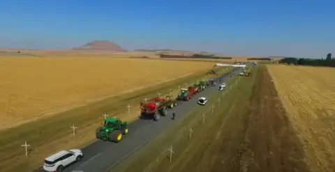 YouTube Rows of white crosses can be seen on either side of a rural road in South Africa. Tractors and cars drive down the middle of the track, with fields on either side .