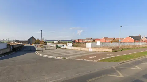 Google A school is on the left-hand side of the image. To the right, there is fenced-off, derelict land. A road runs in front of the site.