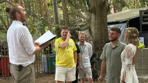George King/BBC Craig Brown and Tara Sweeting are smiling and laughing as they get married in front of a taller man wearing a white shirt with a script in his hand. In the background are two other men, who are casually dressed.