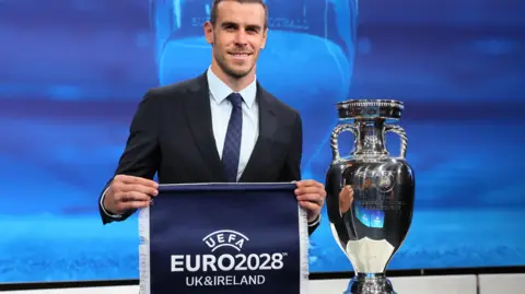 Reuters Former Wales footballer Gareth Bale holds up a blue pennant with the words Uefa Euro 2028 UK and Ireland