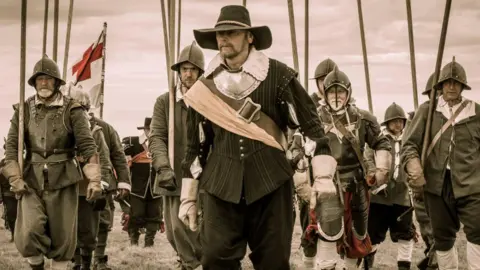 An English Civil Ware enactment, showing an image of several soldiers, wearing historic outfits, armour, holding sticks. They all have hats or helmets on. 