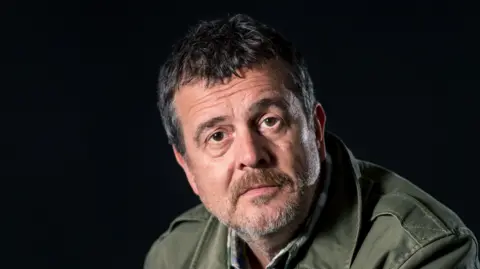 A close-up of a man with dark grey hair looking into the camera. He wears a dark-green jacket and is in front of a dark background.