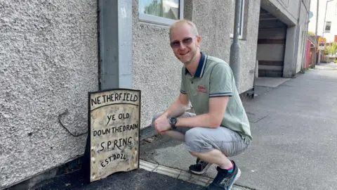 BBC Pip Algar kneeling next to the sign which commemorates the "Netherfield Spring", an area which leaks water often.