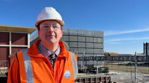 Tom Jackson/BBC Philip Holbourn is standing on the construction site wearing a white hard hat, safety glasses and an orange high viability coat over a shirt and tie. Its is a sunny day with blue skies. 