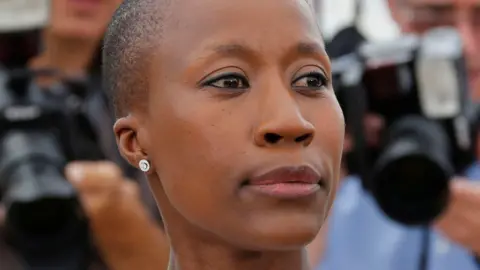 Rokia Traoré has a shaved head and wears diamond stud earrings. The image is a close up of her face and she is looking to her left. In the background there are people with cameras