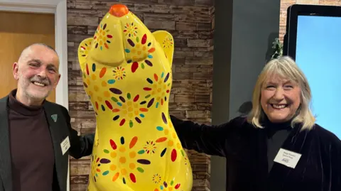 The first rat to be unveiled is titled 'Daisy Rat'. It is a large yellow sculpture of a rat hand painted with multi-coloured dots and petals and an orange nose. A man and a woman are stood either side of the statue, with their arms outstretched and smiling wide.