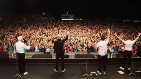 PA Media Four men standing on a stage with their backs to the camera waving to a large crowd facing them