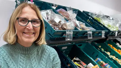 A woman wearing a blue jumper and black frame glasses. She is smiling at the camera. Behind her are rows of food supplies in green boxes 