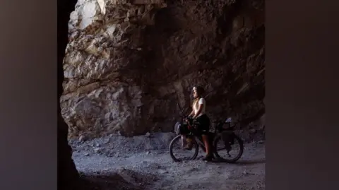 Matilda Pine Matilda in a dark tunnel looks out towards the light while astride her bike. She wears a cycle helmet. 