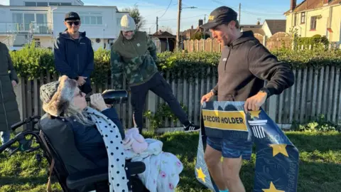 Supplied A man in running gear holding a blue banner that says "world record holder". He is standing in front of a woman who is drinking from a bottle of wine. Two men are smiling behind them.