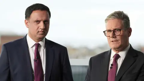 Two  men stand side-by-side posing for a photograph. They are both wearing dark suits, white shirts with ties. 