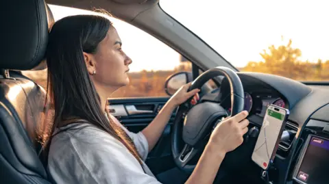 Getty Images A young woman with long, straight, dark hair driving a car and using a hands-free navigator on her smartphone (stock image) 