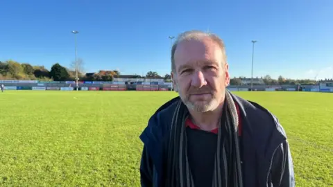 Ian Hopewell with a blue jacket and dark scarf stood at the football pitch.