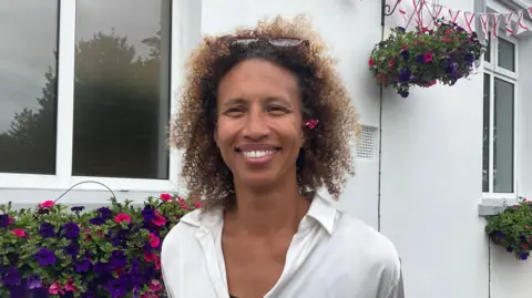 Serena Guthrie smiles are the camera. She has short curly hair with sunglasses on top of her head, and is wearing a white blouse. Behind her is a white building with flowers outside the window sills, and Jersey flag bunting in the top-right corner. 