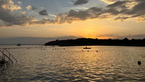 The sun is seen rising over Clevedon Marine Lake at dawn. The water is calm and the sun is lighting up the horizon beyond the pier with bright orange hues. There are some swimmers in the water, visible only as silhouettes.