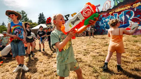 Shindig Festival Three children playing with water guns at Shindig festival. The surrounding grass is brown and scorched from the sun. 