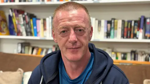 A man with short brown hair, wearing a blue hoody and blue t-shirt, sits on a brown sofa. There are bookshelves behind him, lined with books.