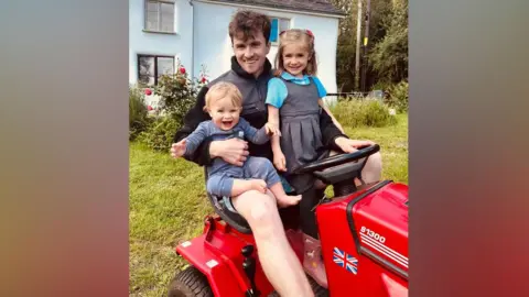 A young dad is sat on a red ride-on lawn mower with his two children in his lap. One is a young blonde boy aged around 16 months, the other is a five-year-old girl with light brown hair in plaits. The girl is wearing her school uniform. 