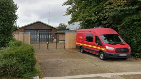Google Google Street view of the old fire station. The site is sealed off with wooden panels and padlocked metal gates. On the driveway in front of the gates is a red fire service crew van.