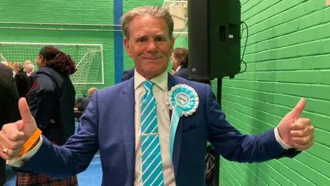 A man wearing a suit and a Reform badge, giving a thumbs up.