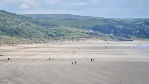 PA Media Woolacombe Beach has just a few walkers, a few with dogs, on a bright day. It is golden sand and the sea is out. The sand dunes are green and there are a couple of houses amidst them.