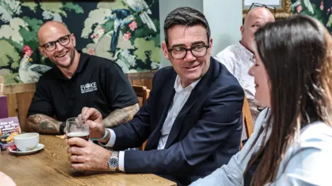 PA Media Andy Burnham at a table in the pub with a man and a woman next to him. He is smiling while listening to the woman talk. The woman has long brunette hair and is wearing a light blue top. The man has a bald head and tattoos on his arms, and he is wearing black framed glasses and a black polo neck shirt. 