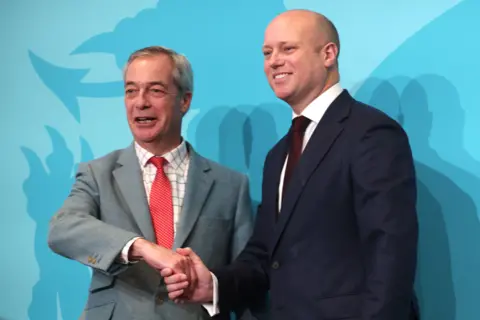 Two men in suits in front of Blue Reform-branded background, shaking hands