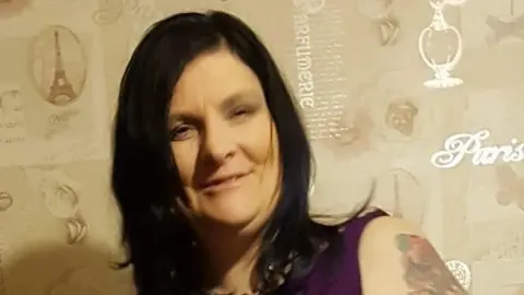 Facebook Ruthanne Jennings is smiling at the camera. She has long dark hair and a tattoo on her left arm and wearing a purple short sleeved top.