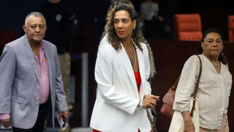 Getty Images Aniella Franco pictured wearing a white blazer over a red outfit. She is pictured in the centre with her father on the left and her mother on the right. 