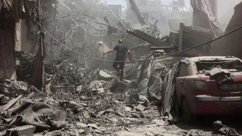 Getty Images A man, walks, his back to the camera, amid grey rubble and a damaged car