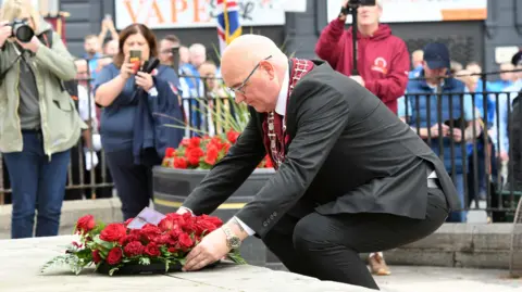 Pacemaker William Walker kneels to lay a wreath of red roses. He is wearing a suit and a maroon sash. There are three people in the background, taking pictures or filming