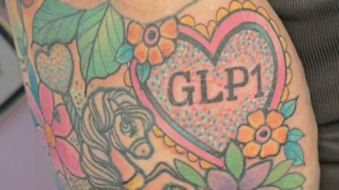 Sian Robinson A sleeve of tattoos, including the lettering GLP1 in a pink and blue heart outline, decorated with yellow flowers and surrounded by other flowers, leaves and a pony.