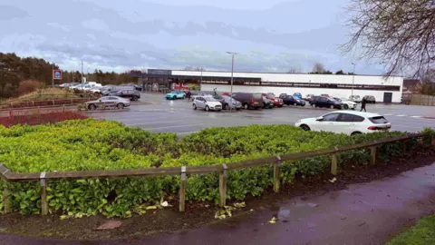 Supermarket Aldi car park in Washington. There are lots of cars in different models and colours parked up in lines outside the white building. There are green bushes surrounding the fenced off perimeter. 