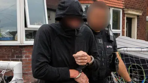 Greater Manchester Police A police officer dressed in black is escorting a handcuffed man wearing all black. They stand in front of a house. Both of their faces have been blurred.