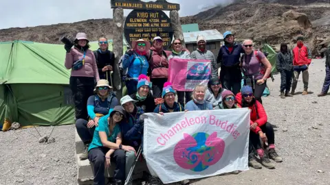 A group of women stand in front of a sign for Kilimanjaro National Park. Several are holding large banners with the words "Chameleon Buddies" written on.