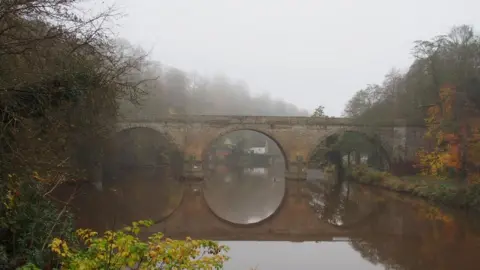 Geoff Dabbs An old stone bridge with three large arches is reflected perfectly in the river beneath it. It is a misty autumnal day with the trees turning orange and yellow.