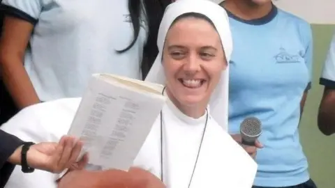 Sr Clare Crockett is sitting , dressed in a habit, while reading from a book being held by one of four young women standing around her