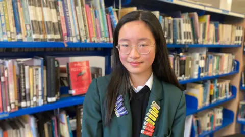 Xiaochu is Chinese name. Xiaochu has black straight hair and circular gold rim glasses. She's wearing her dark green blazer school uniform and has a number of achievement badges on her lapel. Behind her is a row of books in a library setting.