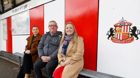 Nexus Councillor Leonard, Ian Gallagher from Nexus, and Roberta Redecke, sit on a metal bench on a metro outdoor platform. Behind them is a red and white panelling, with a football emblem to the right, and a map to the left. A sign to the left, reads 'Stadium of Light'.