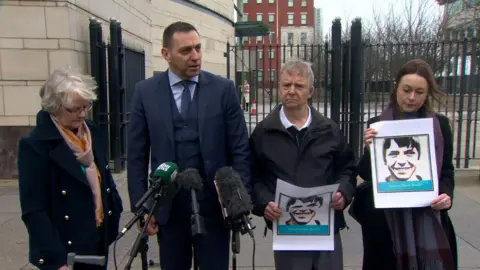 Four people are standing outside a court. Two are holding black and white pictures of a teenage boy. Mics are in front of them. 