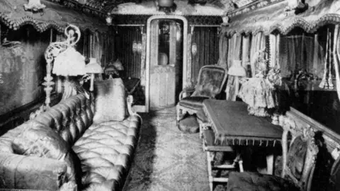 royaltrain.co.uk A photo of the interior of Queen Victoria's carriage on the royal train. There is a long sofa on the left. There are several lamps, two armchairs and decorative curtains on the windows.