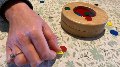 A close-up of a hand playing tiddlywinks, with various coloured counters and a wooden holder on the table-top.