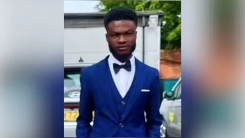 Clinton Antwi with medium-length dark hair and slight beard, wearing a blue jacket and black bow tie. He is standing outside next to a car and in front of a white building.