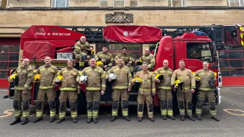 A crew of about a dozen firemen standing proudly in uniform and holding their helmets in front of a fire engine. Four of them are perched on the vehicle. The fire station can be seen in the background