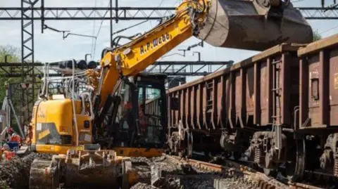 Network Rail Yellow digger scoops up rubble and loads it into carriages to make way for new tracks