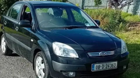 Un Kia Rio Sedán negro, con matrícula 08-D-24363, está aparcado junto al césped. La ventanilla del asiento del conductor está bajada. El vehículo está aparcado sobre grava con césped a la derecha de la imagen y una valla de madera pintada de azul pálido en el borde del marco.