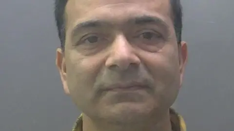 Cambridgeshire Constabulary A custody image of Pramod Dhakal. He has short dark hair and is looking into the camera. He has a yellow and black checked shirt collar.